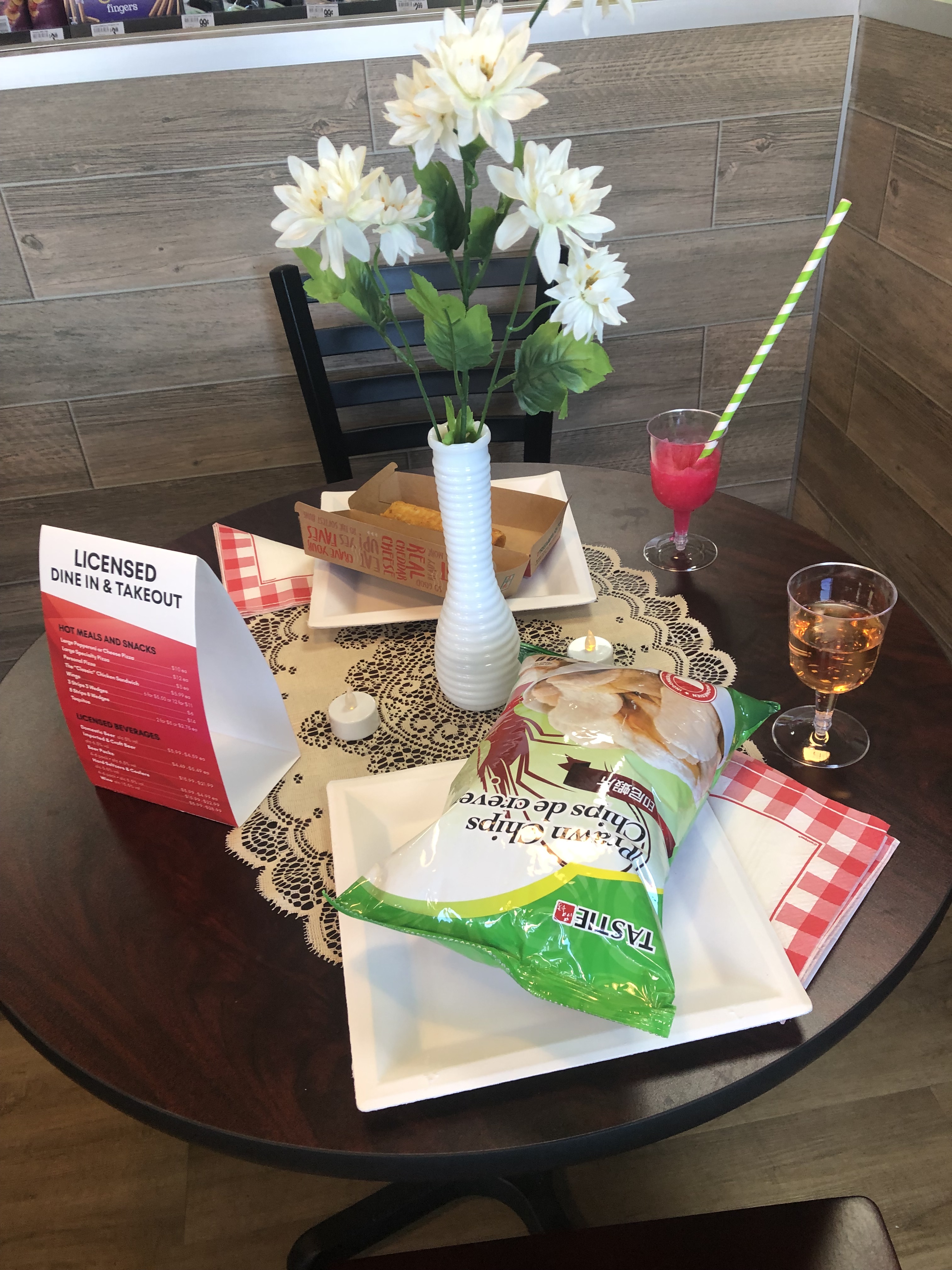 Dining Table with a mini tablecloth and flowers in a vase. A plastic wine cup, Slurpee, taquito, and chips. 